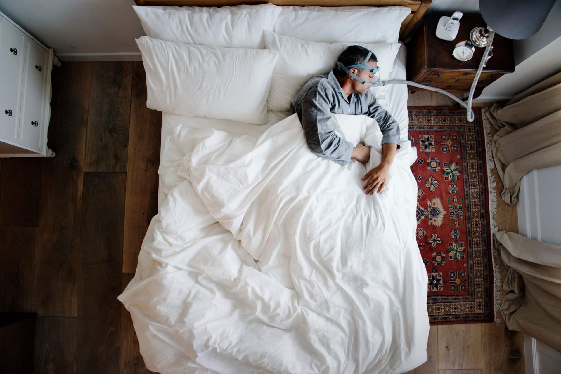 A person sleeps on a bed with white sheets, wearing a CPAP mask. The room has wooden flooring, a bedside table with a lamp and alarm clock, and a patterned red rug. The person is covered with a white comforter.