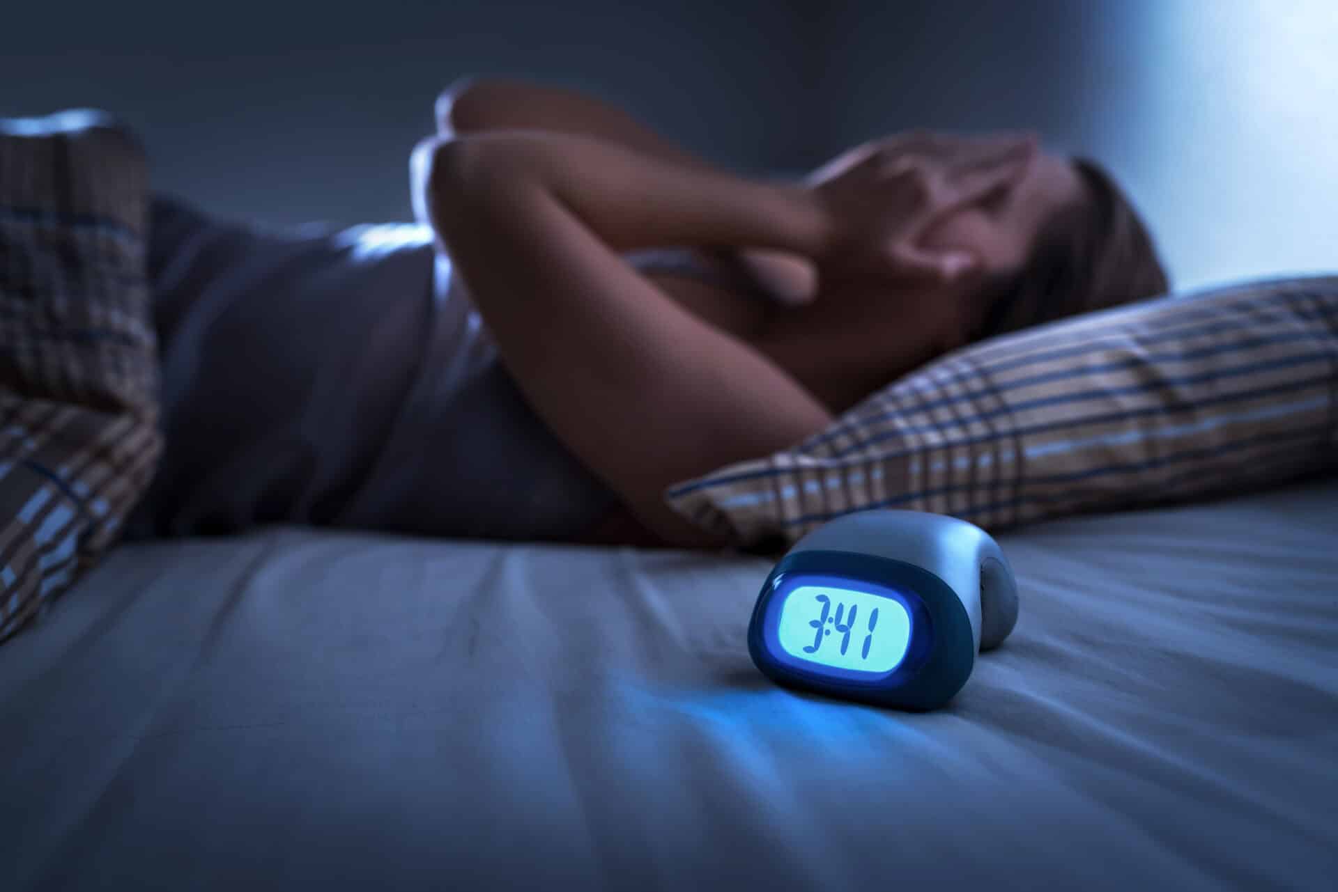 A person lying awake in bed with their hands covering their face. It's dark, and an alarm clock on the bedside shows 3:41 a.m. The scene suggests sleeplessness or insomnia.