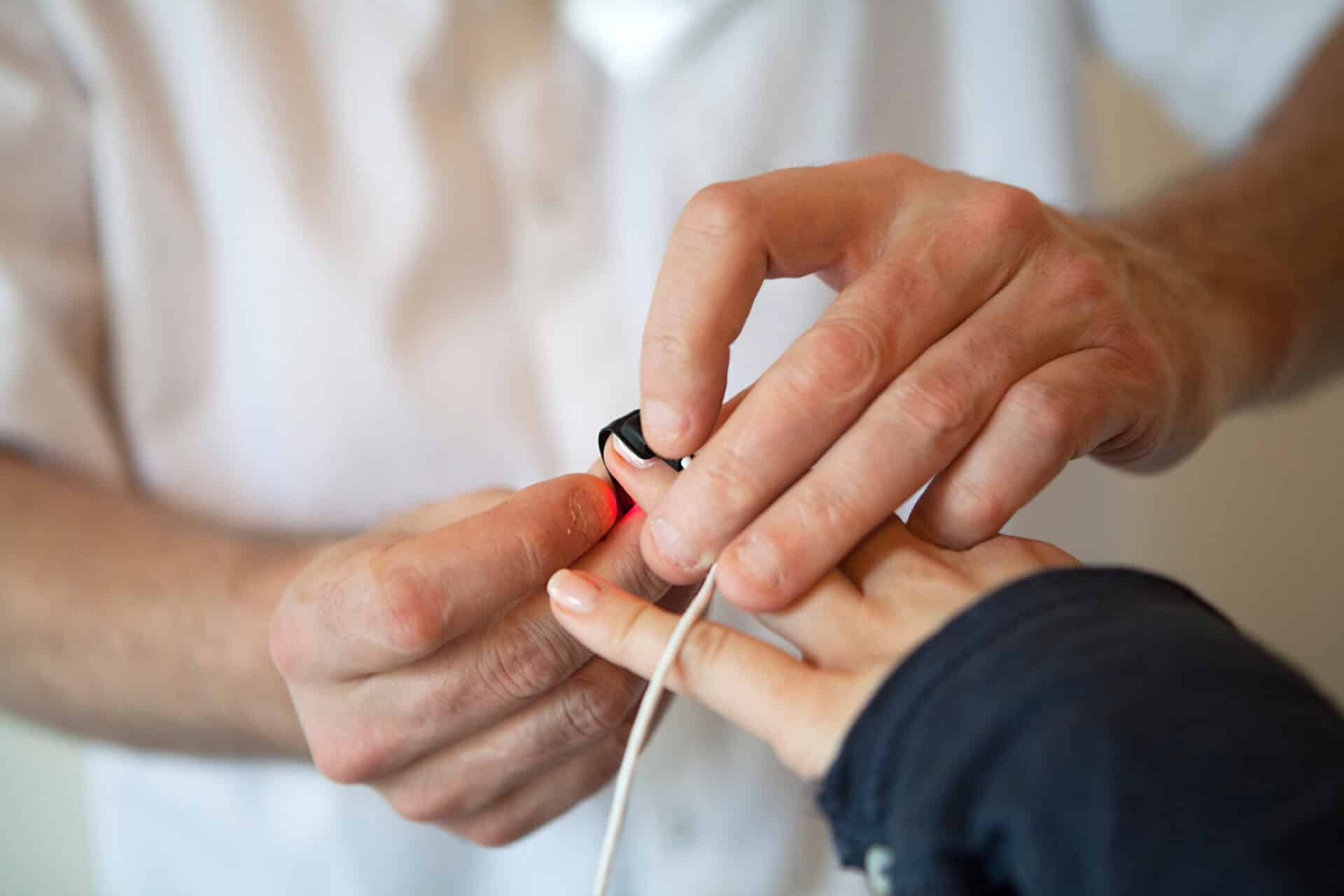 A healthcare professional places a pulse oximeter on a person's finger to measure oxygen levels. The person is wearing a dark jacket.