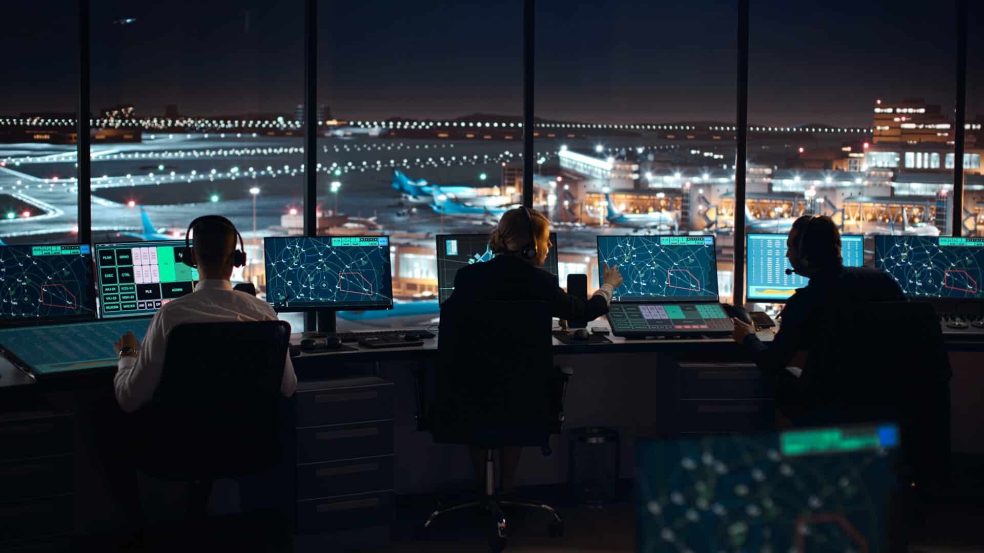 Three air traffic controllers are seated at a row of computer screens in a tower overlooking a busy airport at night. The screens display flight paths and data. The bright airport runway lights are visible through the large windows.