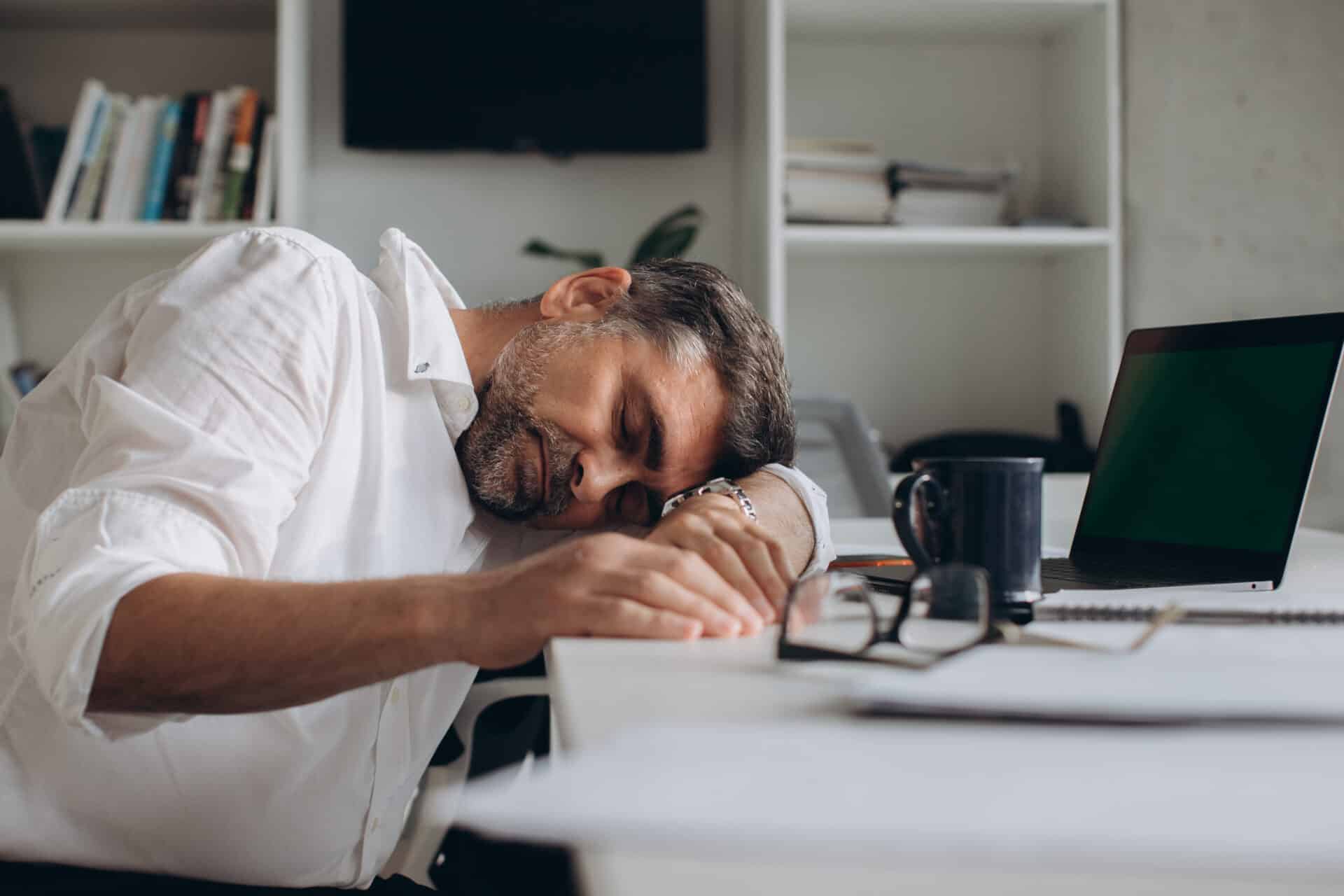 A man in a white shirt sleeps with his head resting on his arm at a desk. In front of him are a pair of glasses, an open laptop, a notebook, and a coffee mug. Shelves with books are in the background.