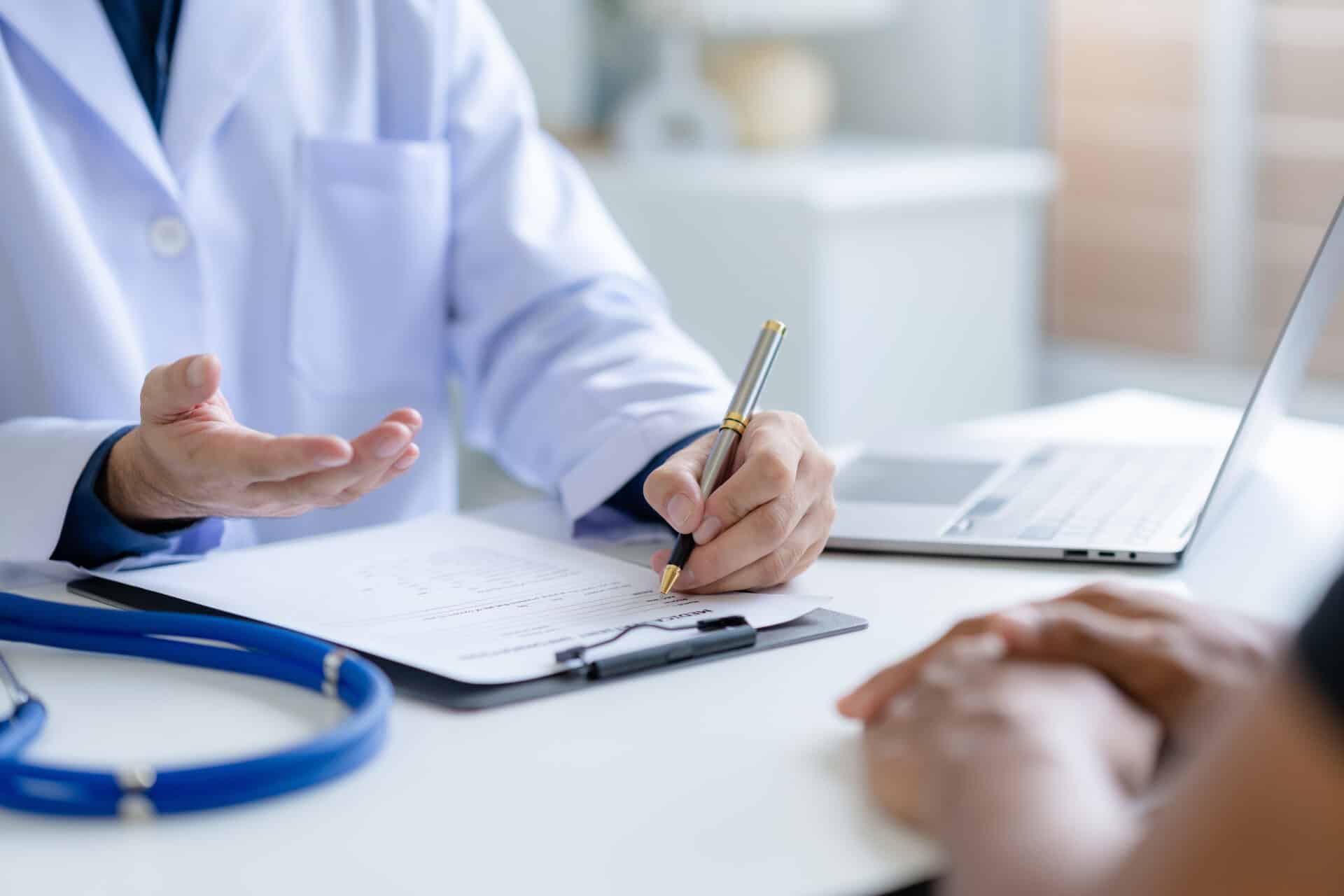 A doctor in a white coat sits at a desk, holding a pen and taking notes on a clipboard. A patient sits across the table. A stethoscope rests on the desk next to an open laptop, suggesting a consultation or medical discussion.