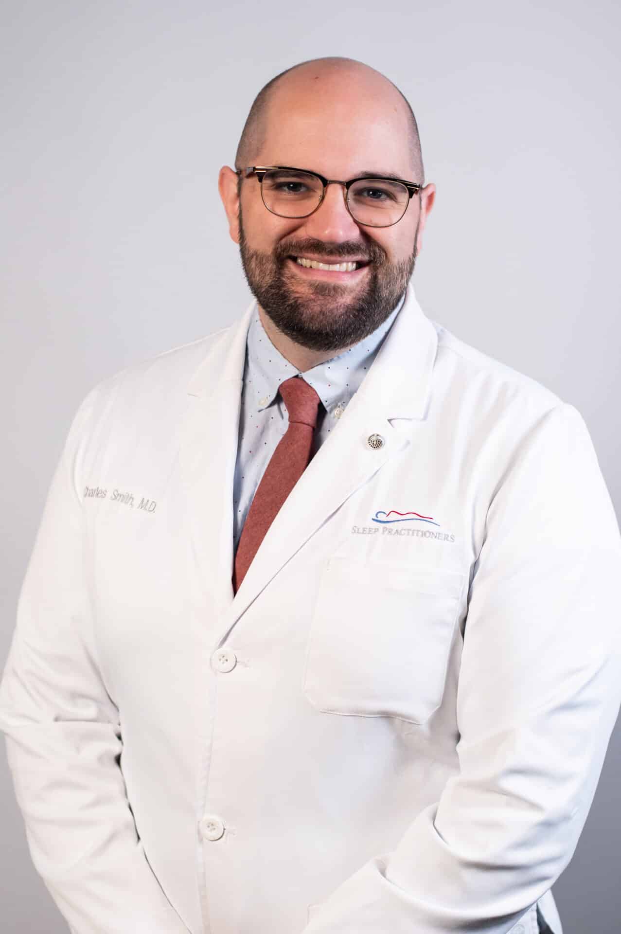 A smiling man with a beard is wearing glasses, a white medical coat, and a red tie against a neutral background. The coat has text and a logo embroidered on it.