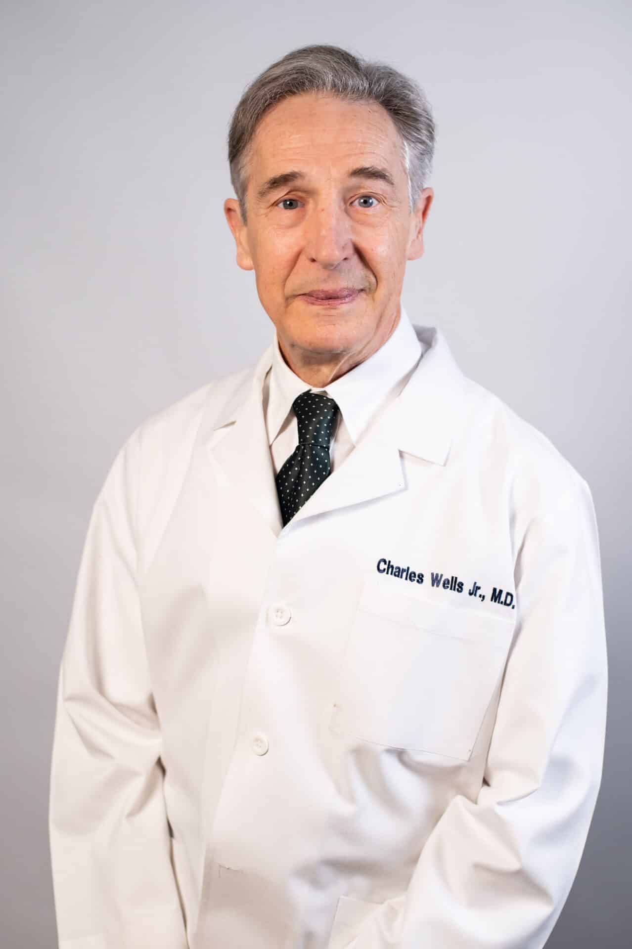 An older man with short gray hair is wearing a white doctor's coat, a white shirt, and a dark tie. He is standing against a plain background and looking at the camera.