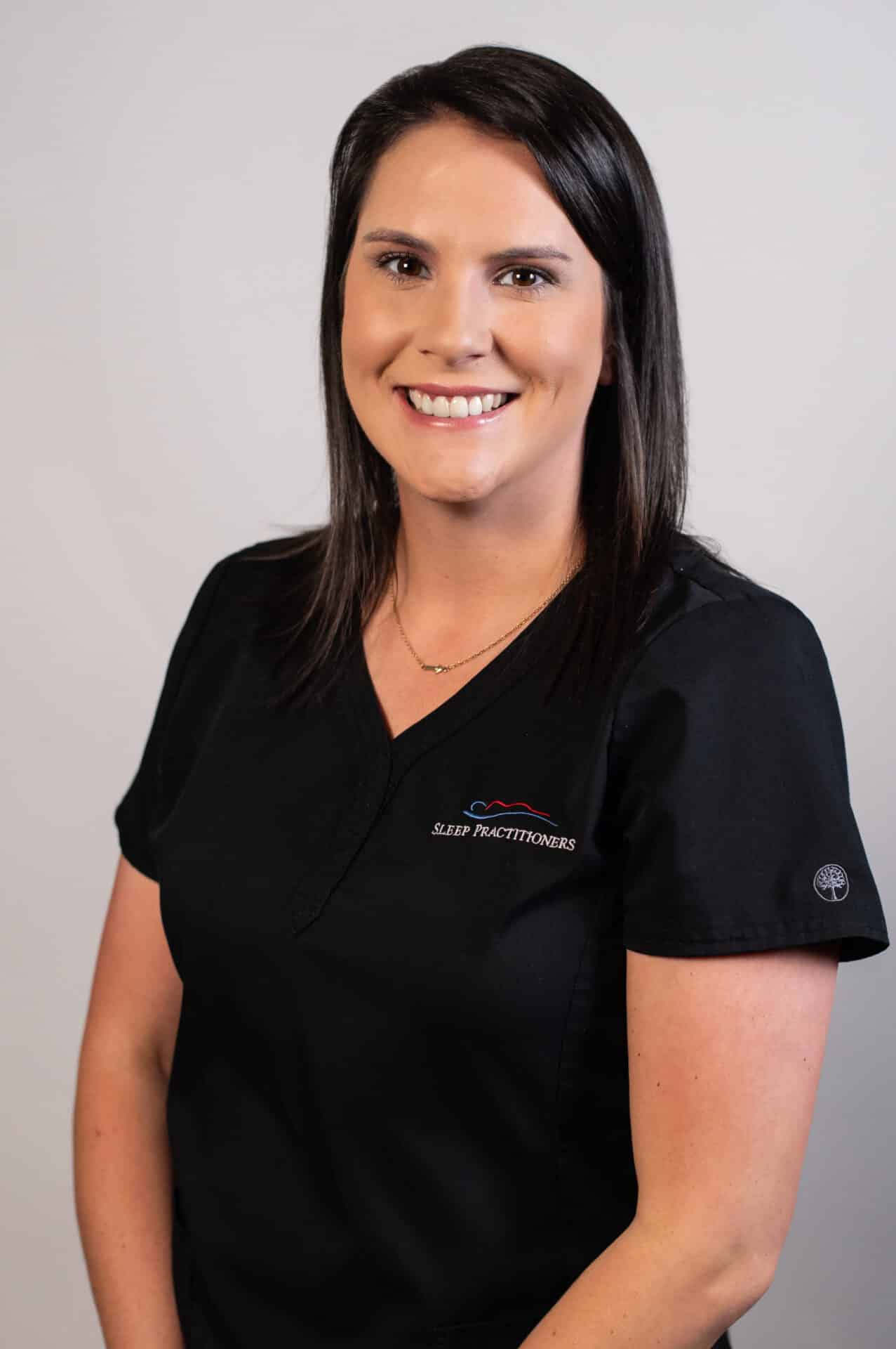 A woman with shoulder-length dark hair smiles while wearing a black scrub top with a Sleep Practitioners logo. She stands against a plain light gray background.