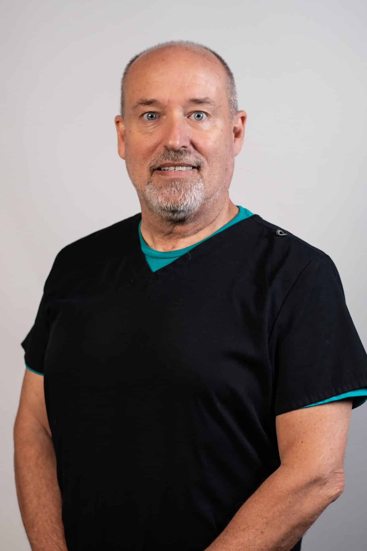 A man wearing a black shirt with a teal undershirt is standing in front of a plain background. He has short hair, a beard, and is looking directly at the camera with a neutral expression.