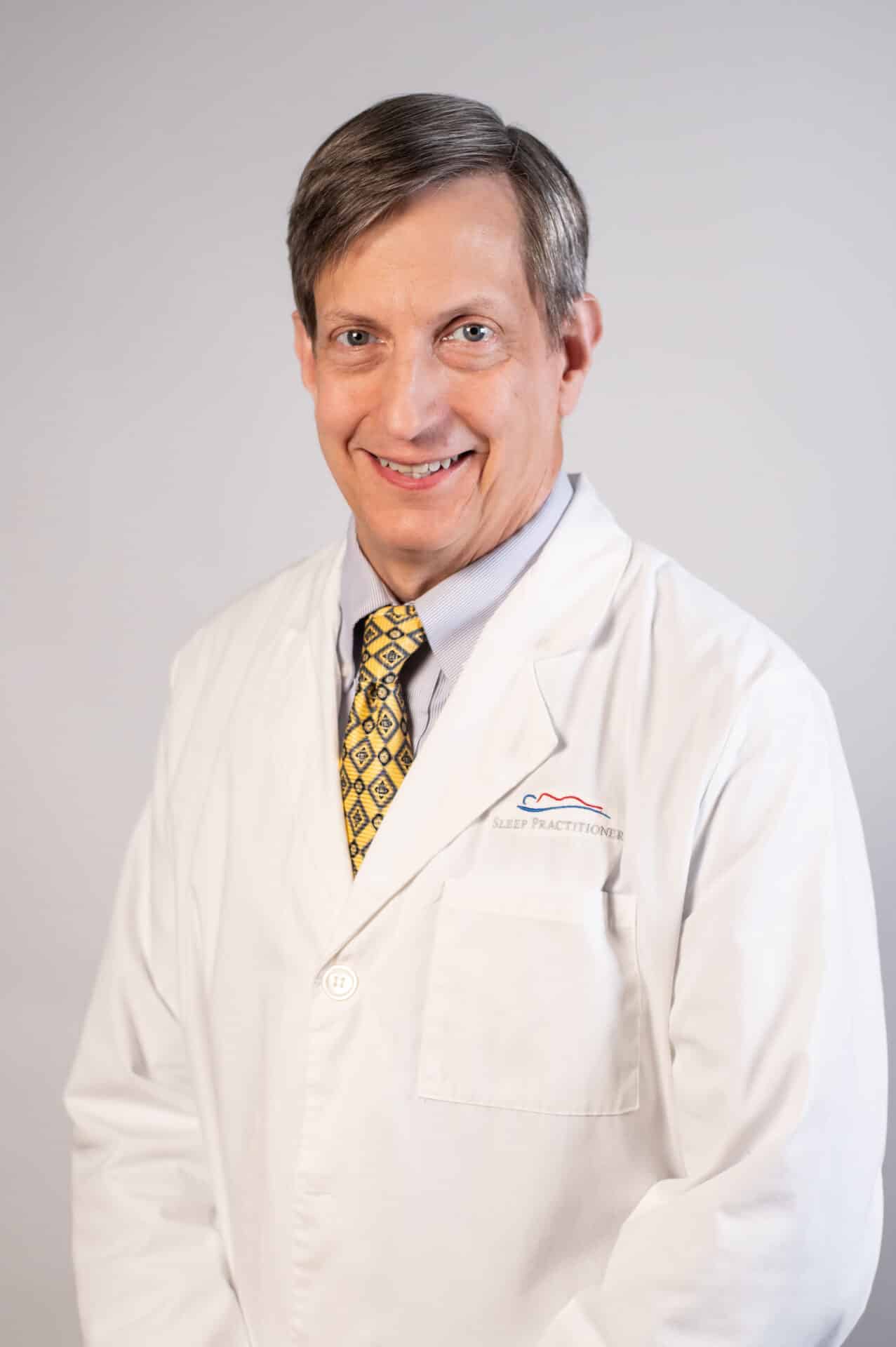 A male doctor smiling, wearing a white lab coat and a yellow patterned tie, standing against a plain background.