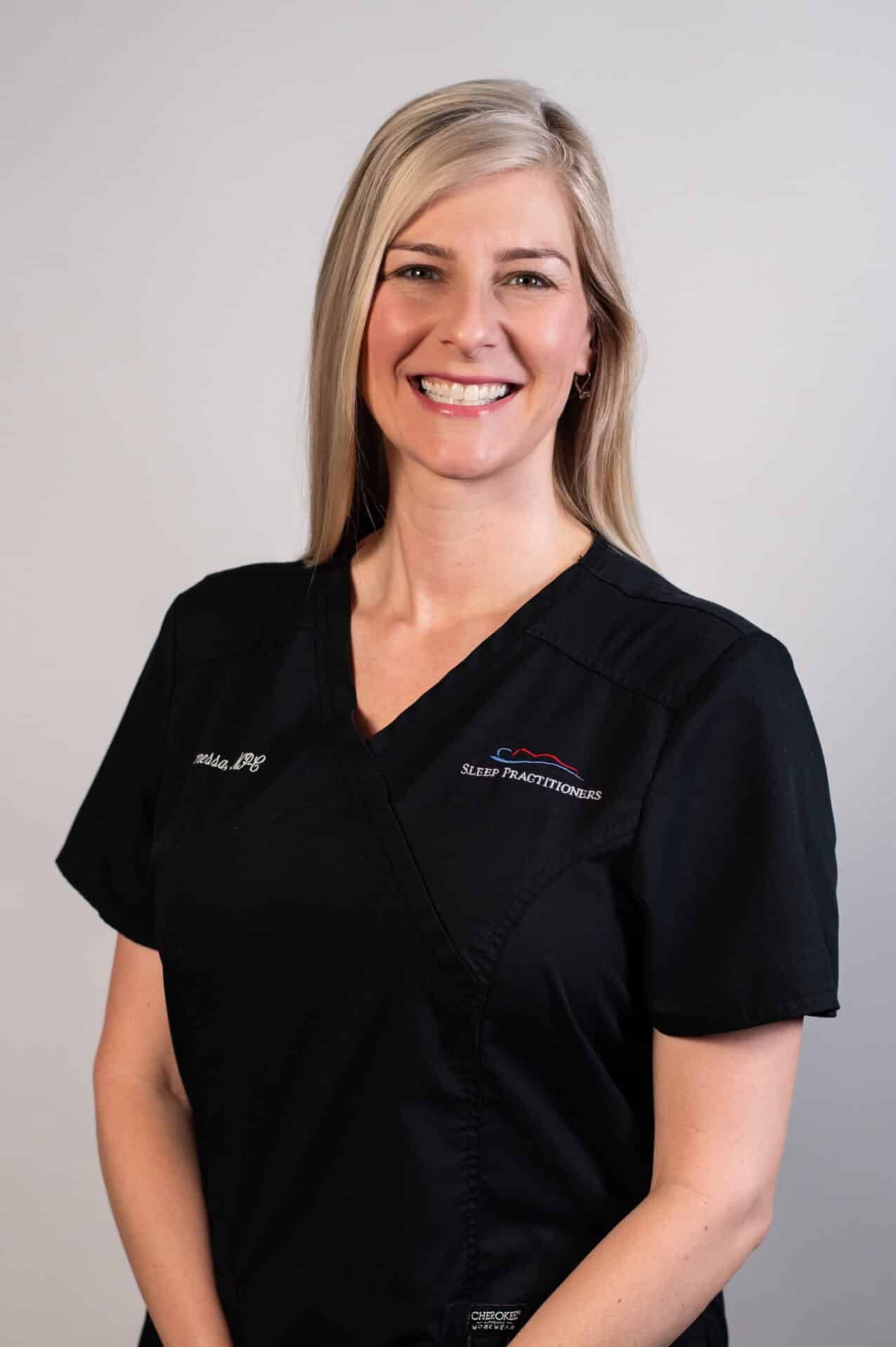 A smiling person wearing black medical scrubs stands against a neutral background. Their name and title are embroidered on their uniform, and their long blonde hair is worn down.