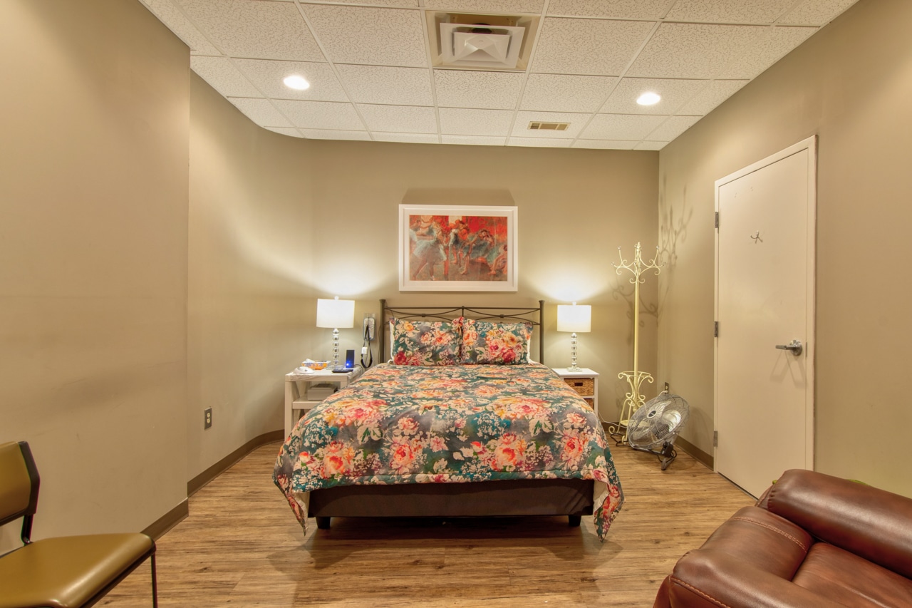 Cozy room with a floral-patterned bed, two bedside tables with lamps, and artwork above the bed. A coat rack stands by a closed door, and a leather chair is in the foreground. The room has warm lighting and wooden floors.