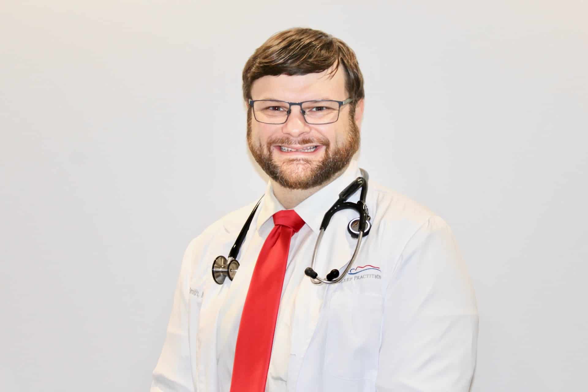 A male medical professional with a beard and glasses is smiling at the camera. He is wearing a white coat, red tie, and has a stethoscope around his neck, standing against a plain white background.